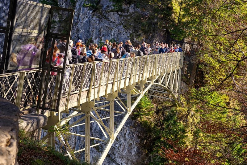 People standing on the Marienbrücke in a forest area surrounded by mountains, autumn atmosphere and sunshine, near Neuschwanstein Castle, near Füssen, Ostallgäu, Allgäu, Bavaria, Germany