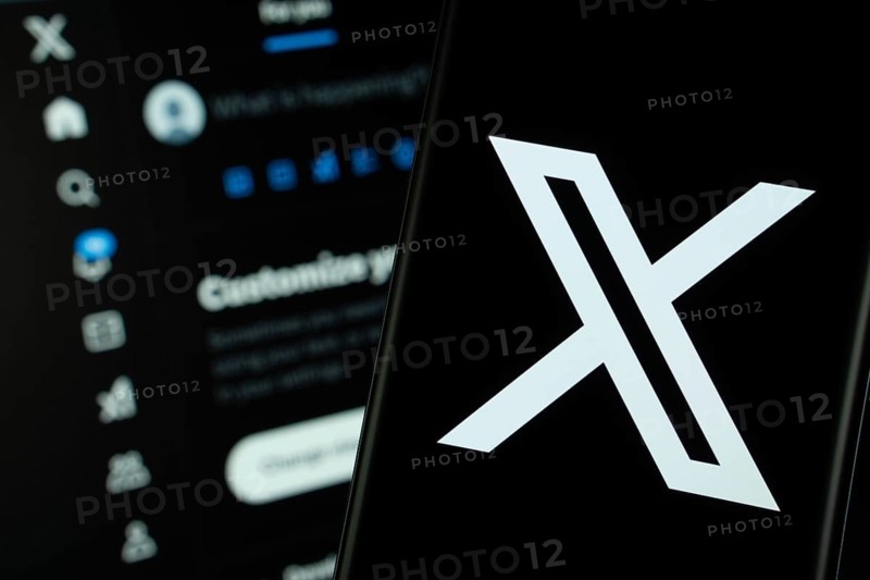 X (twitter) logo is seen on a smartphone, with the bluesky website visible on a computer screen in the background