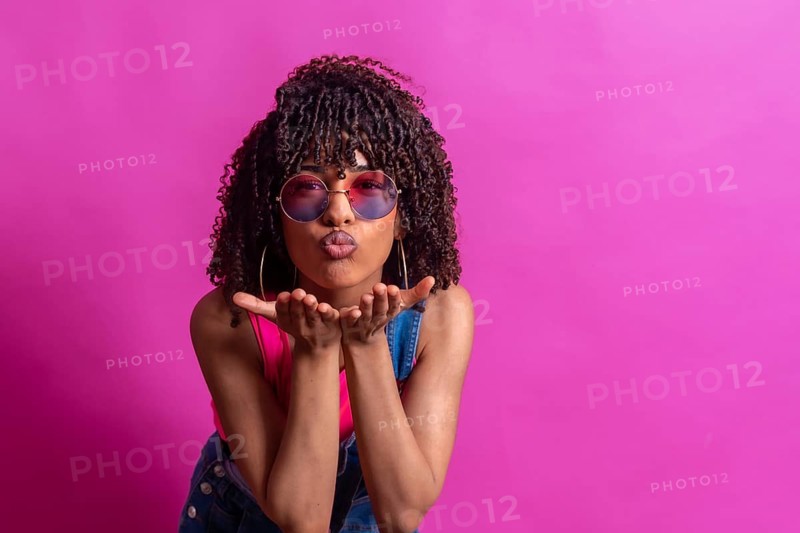 Fashionable young woman blowing a kiss while wearing sunglasses, set against a vibrant pink backdrop