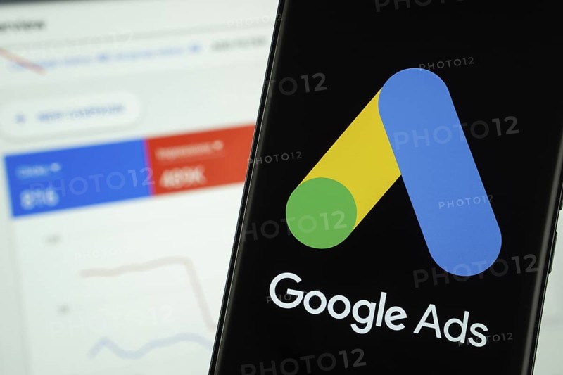 Google Ads logo is displayed on a smartphone screen, with a stock graph webpage visible on a computer screen in the background