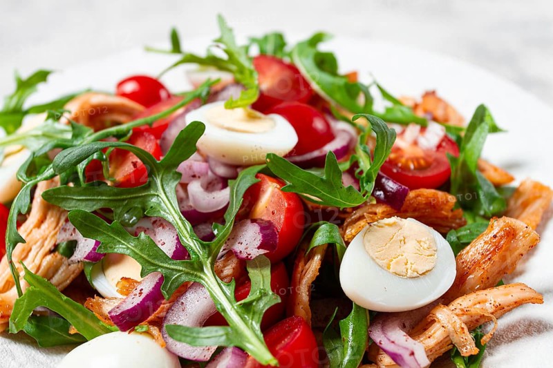 A warm salad with fried chicken fillet, arugula and cherry tomatoes, and boiled quail eggs, on a white plate, no people