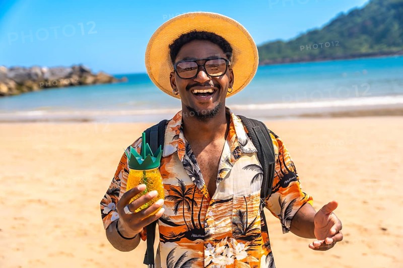 Young man smiling while holding a refreshing tropical drink on a sunny beach, fully enjoying his summer vacation getaway
