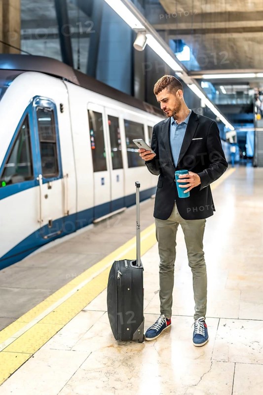 Businessman standing on subway platform, using smartphone, holding coffee, waiting for train with his trolley