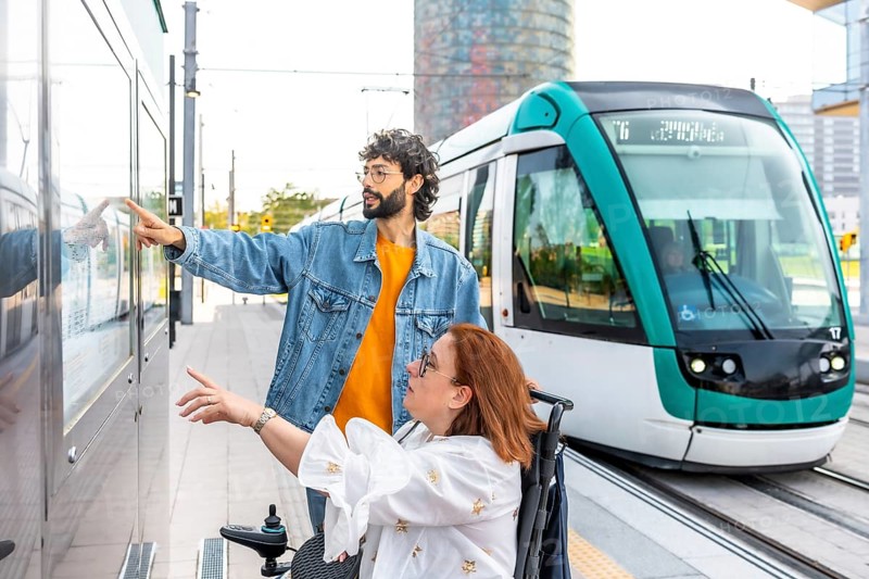 Young man assists a woman in a wheelchair, pointing at the tram schedule at a modern station, ensuring accessible public transportation