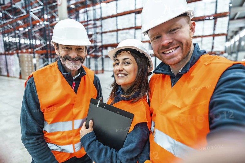 Warehouse workers wearing safety vests and hard hats are smiling and taking a selfie in a logistics center