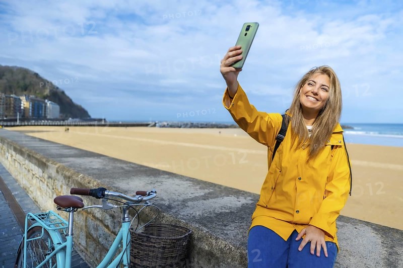 Young woman wearing yellow raincoat taking selfie with smartphone at la zurriola beach in san sebastian, spain, with her bicycle parked nearby