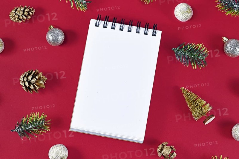 Empty notebook surrounded by seasonal decoration like golden fir cones, fir branches and Christmas tree baubles on dark burgundy background
