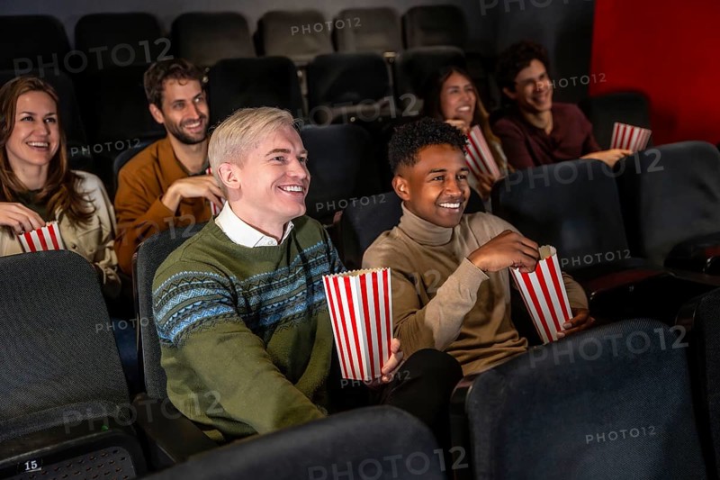 Diverse group of friends laughing and eating popcorn while watching a movie at the cinema