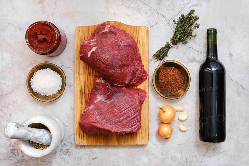 Raw cuts of beef on a wooden board alongside garlic, onions, herbs, spices, salt, tomato paste, and a bottle of red wine, ingredients for beef bourguignon preparation