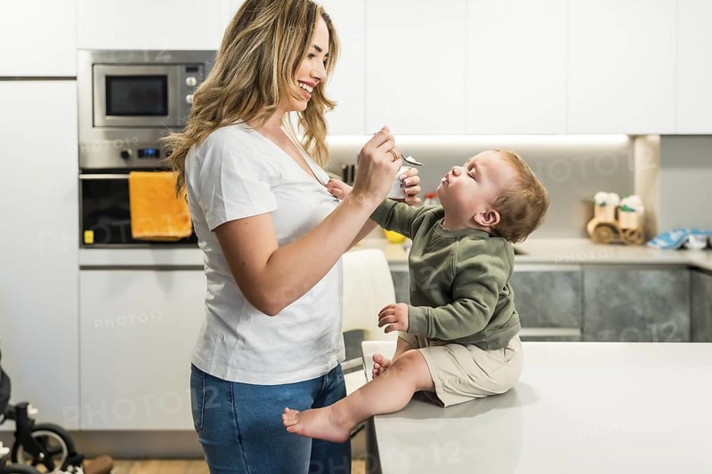 Smiling mother joyfully feeding her baby son while sitting on the kitchen counter, creating a warm moment of love and connection at home