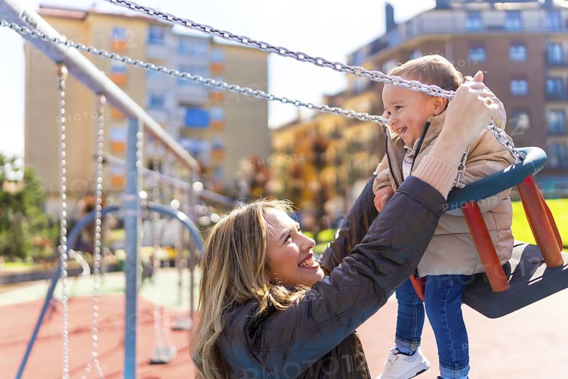 Mother enjoying playful moments with her baby boy, lifting him high on a swing in a vibrant playground under the sunny sky