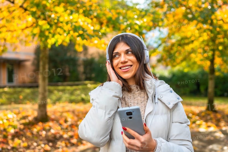 Caucasian young woman enjoying music using mobile phone and headphones standing a city park in autumn