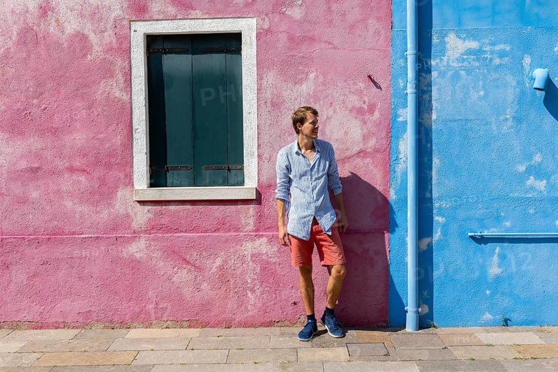 Young man leaning against a house wall, pink and blue house facade with window, colourful houses on the island of Burano, Venice, Veneto, Italy