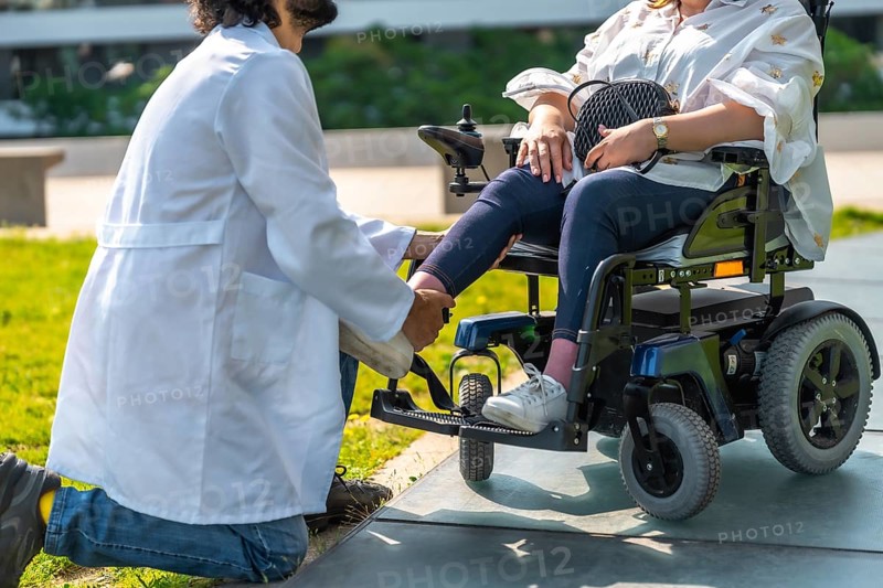 Doctor adjusting the footrest of a patient sitting in an electric wheelchair, providing personalized care and support in a park setting