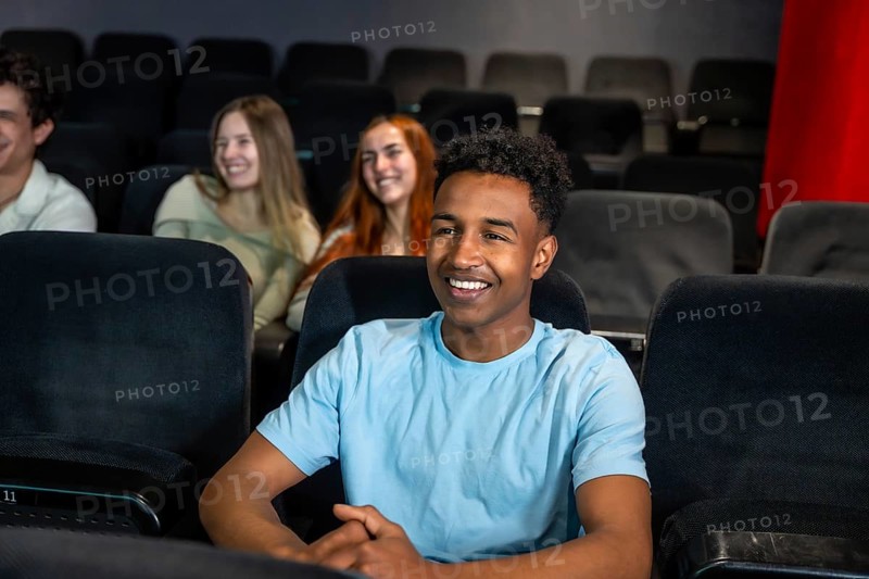 Young man smiles while watching a movie in a cinema, surrounded by his friends, enjoying a shared entertainment experience