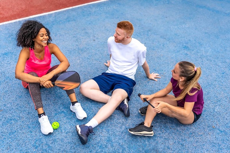 Top view photo of a multi-ethnic group of friends sitting on pickelball court chatting relaxed after play