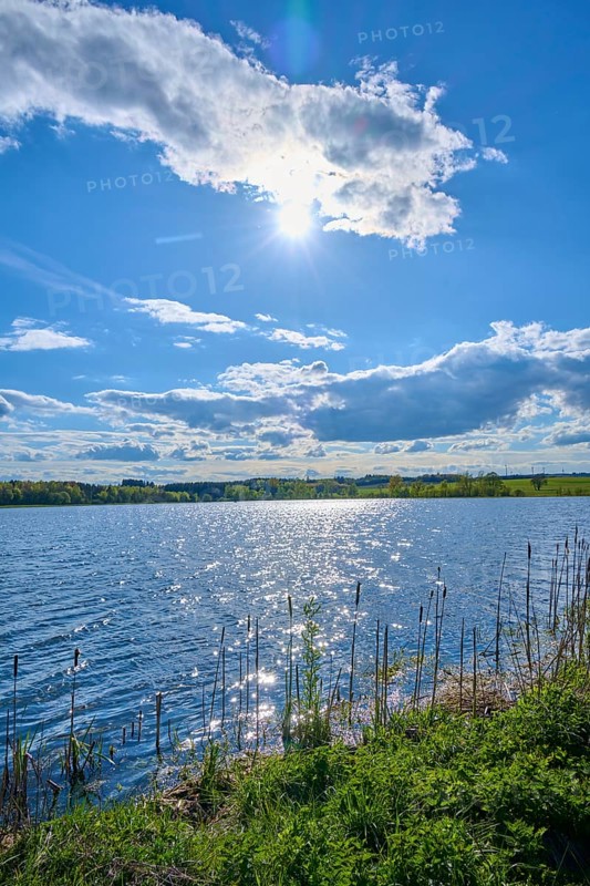 Ober Moosersee surrounded by green fields and trees under a partly cloudy blue sky, spring, Freiensteinau, Vogelsberg, Hesse, Germany