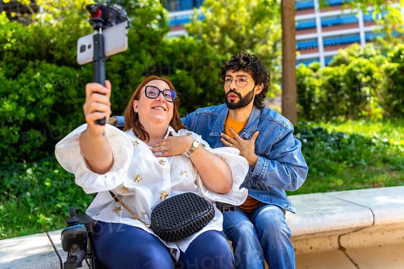 Two content creators sitting on a bench in a park, filming a video blog using a smartphone and gimbal