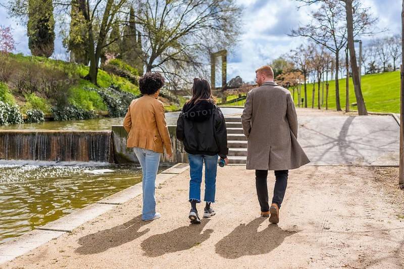 Three friends walking away from camera on path by pond with small waterfall in sunny public park