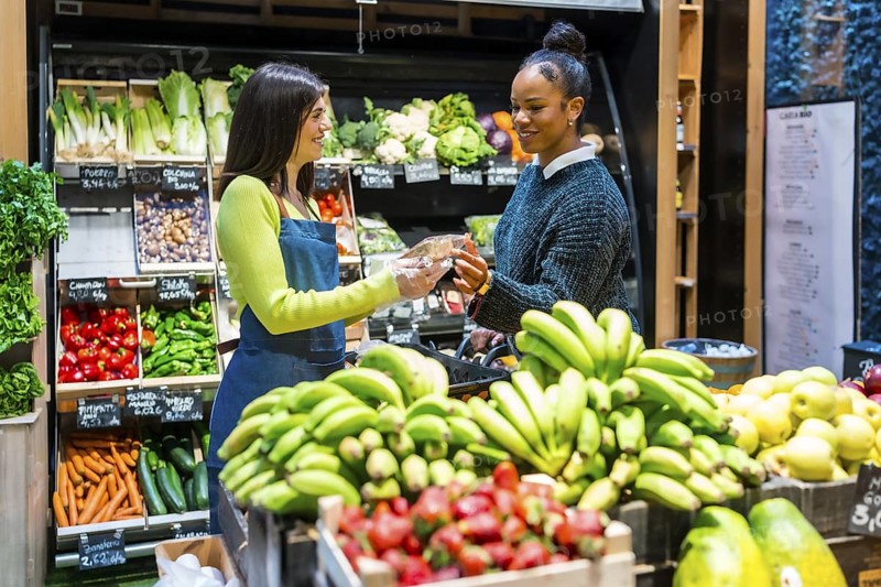Friendly saleswoman handing a packaged product to a customer in a supermarket with shelves full of fresh fruits and vegetables