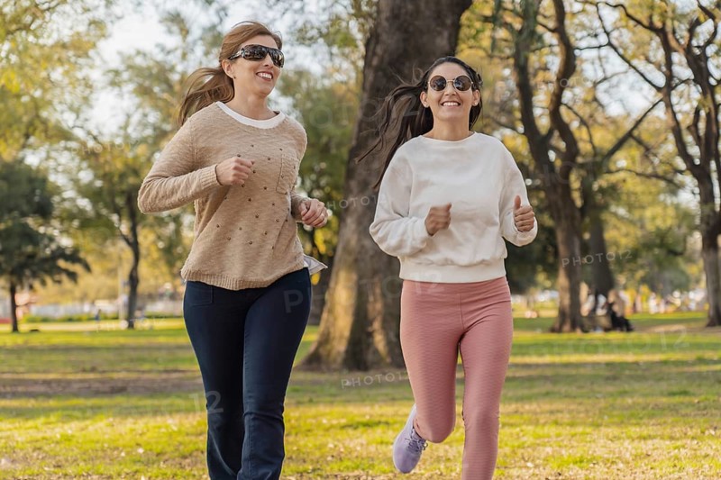 Two women jog side by side in a sunny park, smiling and enjoying the fresh air. Dressed casually in sweaters, they embrace a healthy lifestyle, surrounded by trees and green scenery
