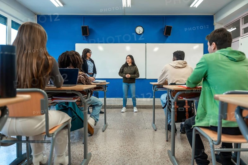 Teenager girl giving a presentation in a classroom while teacher and classmates listen to her