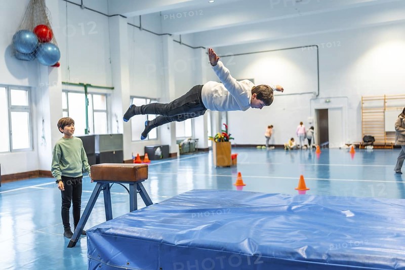 Child practicing vaulting in a school gym, watched by another student and supervised by a teacher, learning gymnastics and improving motor skills