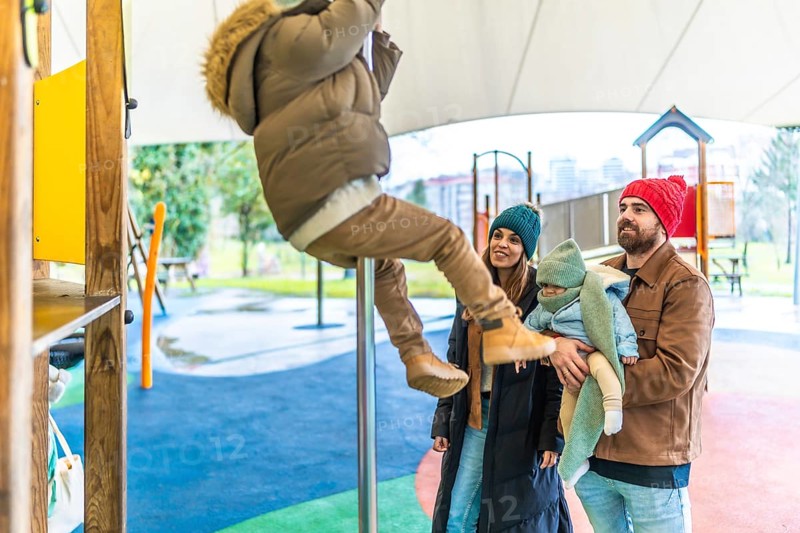Young boy climbing a pole at the playground while his parents and baby sibling watch him, enjoying a winter day outdoors