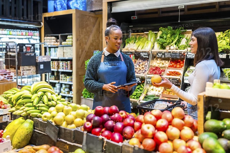 Supermarket employee using digital tablet assisting customer choosing grapefruit in organic supermarket with fresh produce