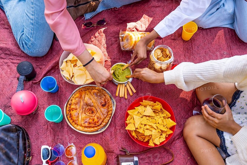 Group of friends having a picnic, sharing snacks like chips, guacamole, pie, and crackers, with drinks on a red blanket
