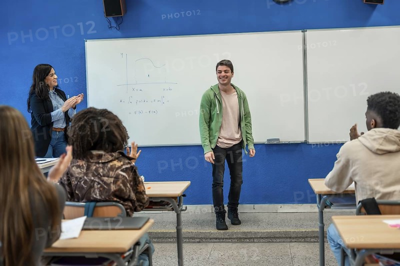 Happy college student explaining lesson at whiteboard while professor and classmates applaud him in modern classroom