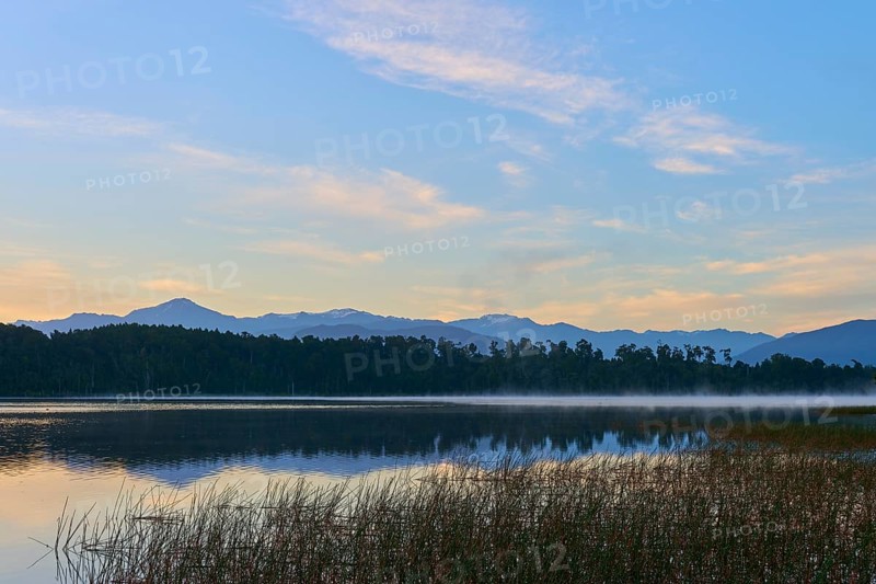 A still lake with a misty water surface and mountains in the background in the early morning, Lake Mahinapua, Ruatapu, South Island, New Zealand
