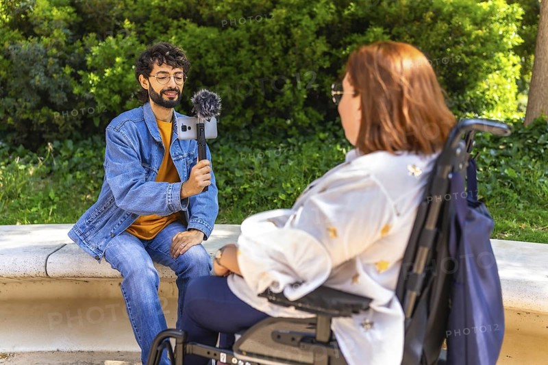 Content creator interviewing a businesswoman in a wheelchair using a smartphone and microphone in a park
