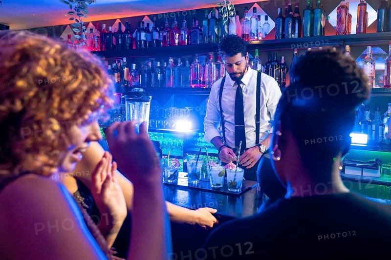 Professional bartender preparing cocktails for cheerful female clients in a nightclub, mixing drinks and creating a vibrant atmosphere