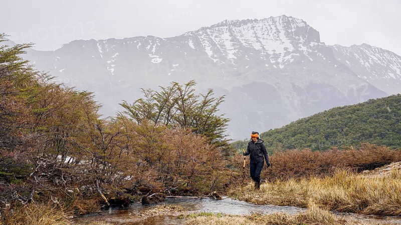 Woman hikes through grassland, Glaciar Vinciguerra, Ushuaia, Tierra del Fuego Province, Argentina, Ein Wanderer bewegt sich entlang eines Flusses vor einer Hintergrundkulisse aus Bergen und Sträuchern, Argentinien