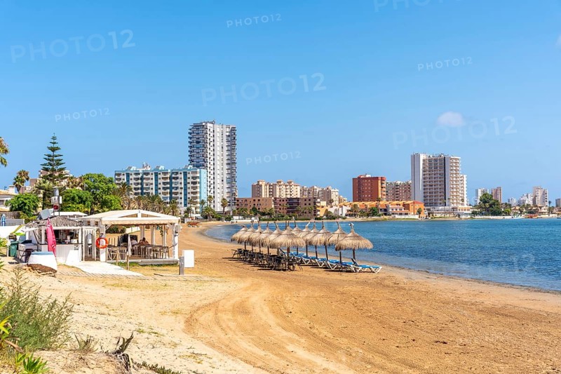 Straw umbrellas and beach chairs sit on the sandy shore of la manga, with resort hotels and apartments rising in the background, Playa de Cavanna