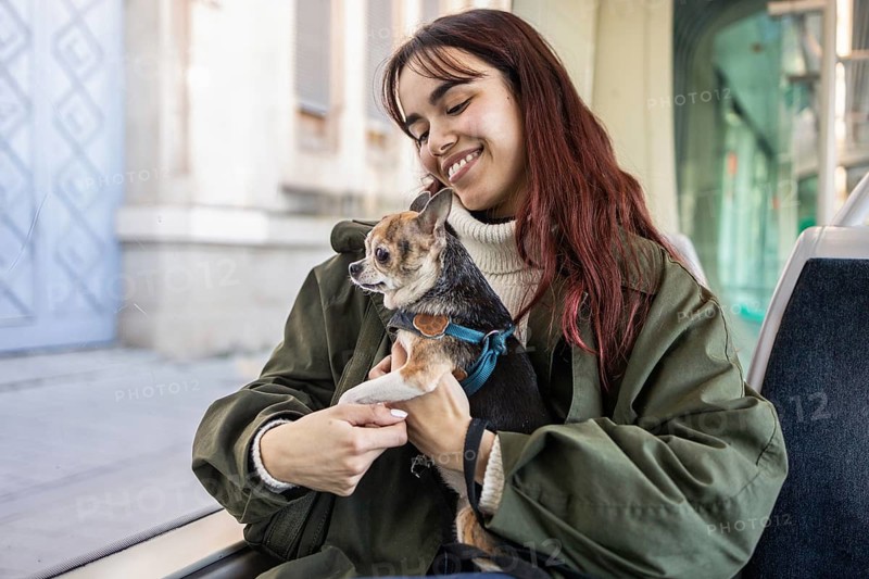 A young woman smiles warmly while holding her small chihuahua on public transport. She's wearing a green coat, and the scene reflects joy and companionship