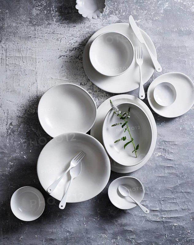 Top view of an elegant arrangement of white ceramic dishes and utensils on a textured grey background. Includes plates, bowls, and a small green sprig as an accent