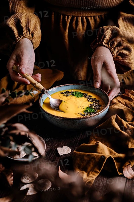 A woman's hands hold a rustic bowl of creamy pumpkin soup garnished with fresh herbs and seeds. The warm setting evokes a cozy autumnal meal prepared with care