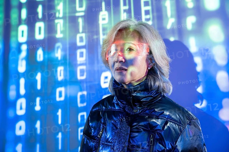 An elderly Asian woman is immersed in the metaverse, wearing futuristic eyewear. The digital background is filled with glowing binary code, symbolizing technology and innovation