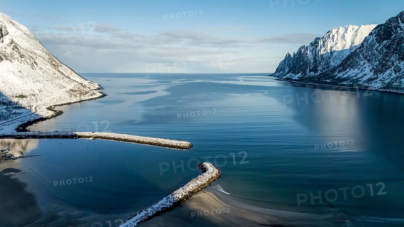 An aerial view showcasing a tranquil Nordic landscape, featuring snow-capped mountains and calm waters under a clear blue sky. The serene scene captures natural beauty, Skaland, Troms, Norway