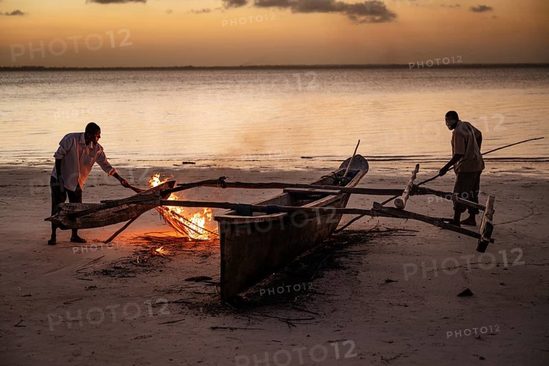 MICHAMVI KAE, ZANZIBAR - OCTOBER 17, 2025: Two local men use a small fire on a sandy beach at twilight to repair or seal a traditional wooden outrigger boat