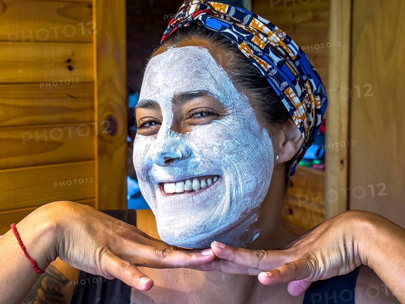 A woman smiling with a white face mask applied, posing playfully in a cozy wooden cabin in Victoria, Australia. Her head is wrapped in a colorful scarf, creating a relaxed, joyful atmosphere