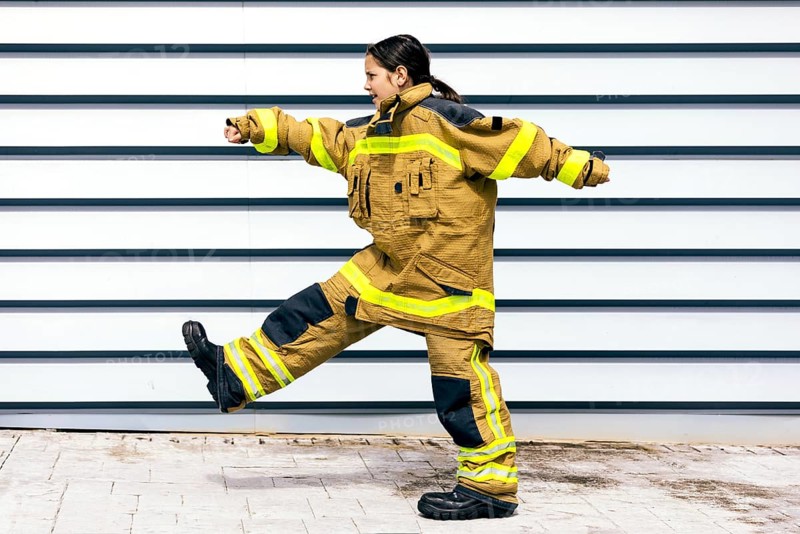 A young girl energetically wearing a firefighter suit, showcasing determination and strength. Her confidence and bravery are evident as she poses against a striped background