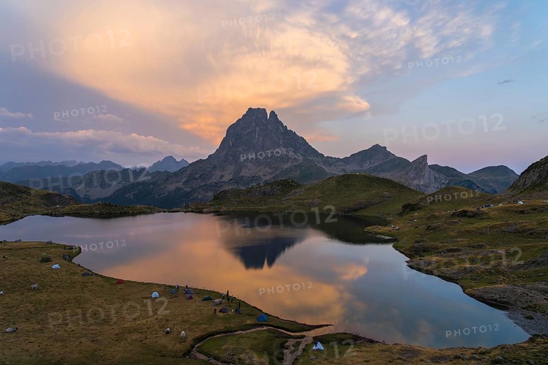 Stunning view of Midi d'Ossau in the French Pyrenees with a serene lake mirroring a colorful sunset Green hills surround the peaceful camping area, creating a tranquil scene
