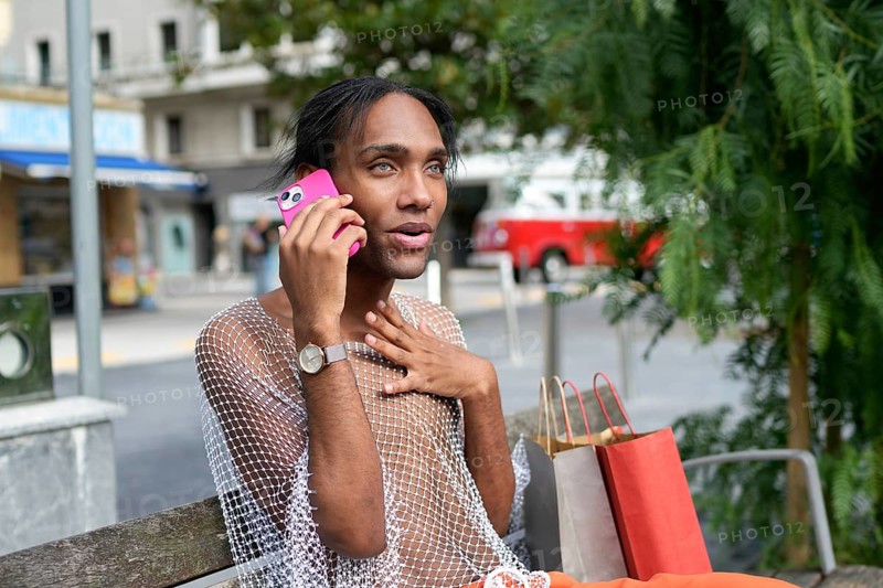 A stylish man in a mesh top converses on a pink phone, seated on a bench in an urban environment, with shopping bags nearby. Capturing pride and self-expression
