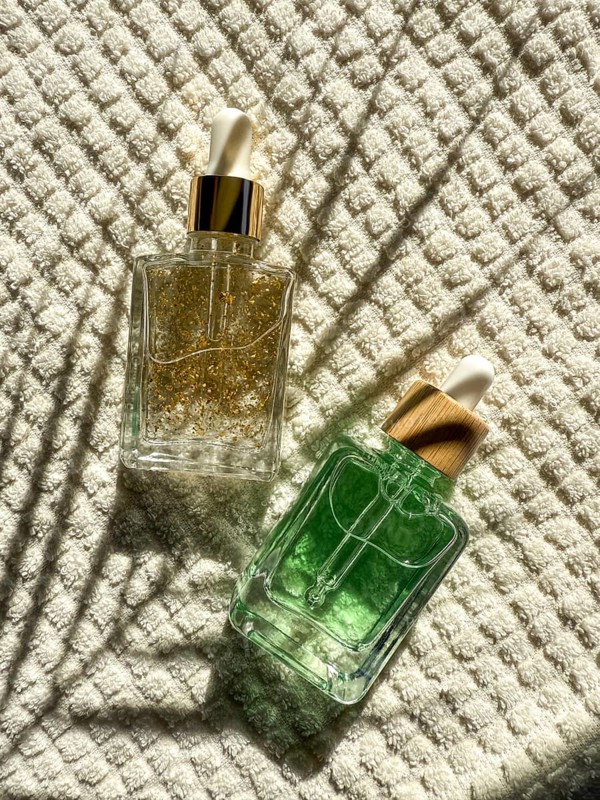 Two elegant dropper cosmetic bottles with gold and green serums lie on textured white fabric int he bathroom. Natural light casts artistic shadows, creating an appealing and luxurious aesthetic