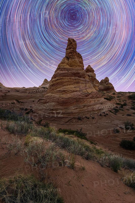 Stunning star trails swirl above a striking desert rock formation, creating a mesmerizing night landscape in Coyote Buttes, Arizona. The scene captures the beauty of nature and time