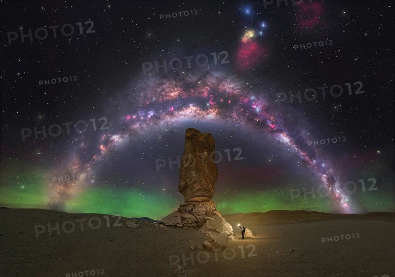 A lone figure stands beneath a towering rock formation, illuminated by the colorful arc of the Milky Way and a subtle aurora, capturing the awe of the cosmic night sky