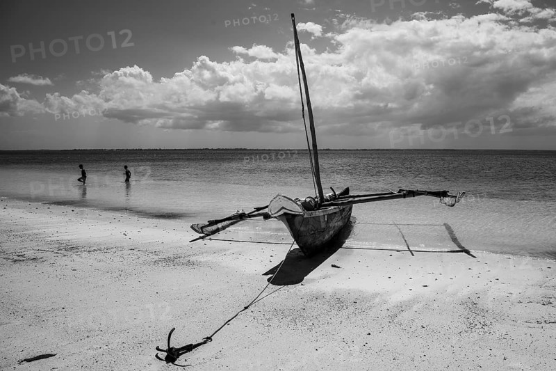 Black and white photograph of a traditional wooden dhow sailboat anchored on a sandy beach under a cloudy sky in Zanzibar, Tanzania
